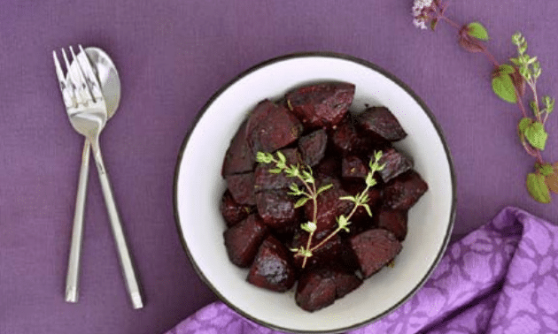 Roasted beets on a white plate with a sprg of rosemary on a purple tablecloth and a fork and knife