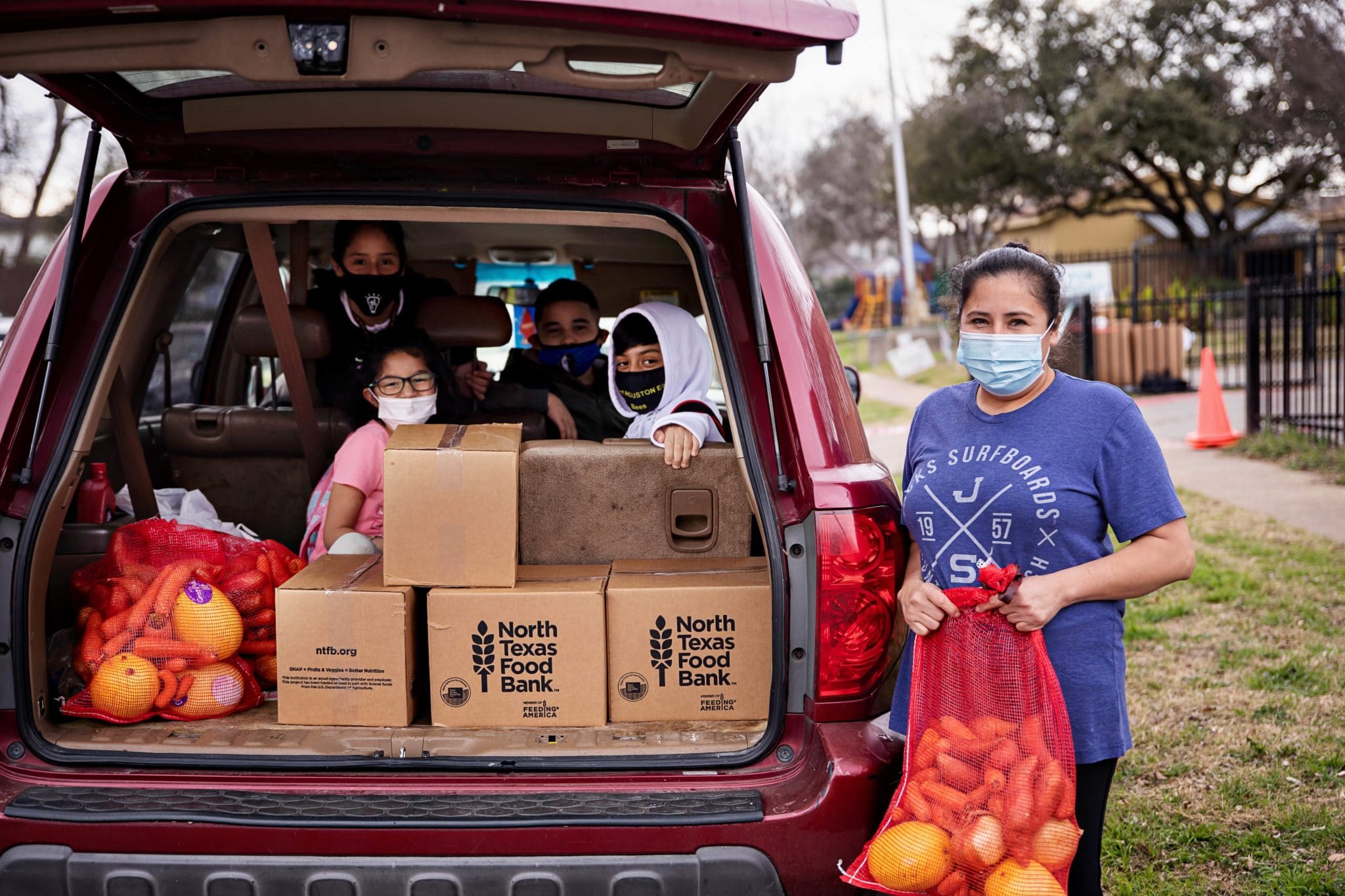 Mother holding a bag of produce next to a car with her children and North Texas Food Bank boxes