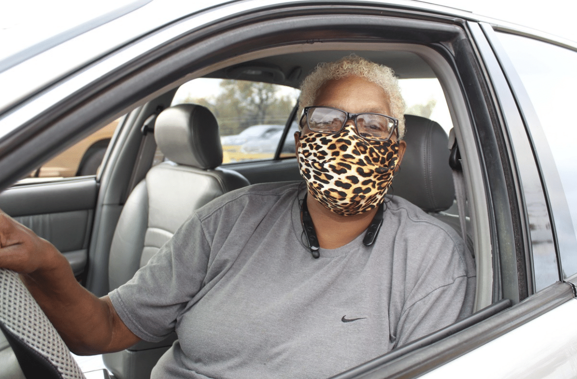 Older black woman wearing a cheeta print mask sitting in the drivers seat of a car