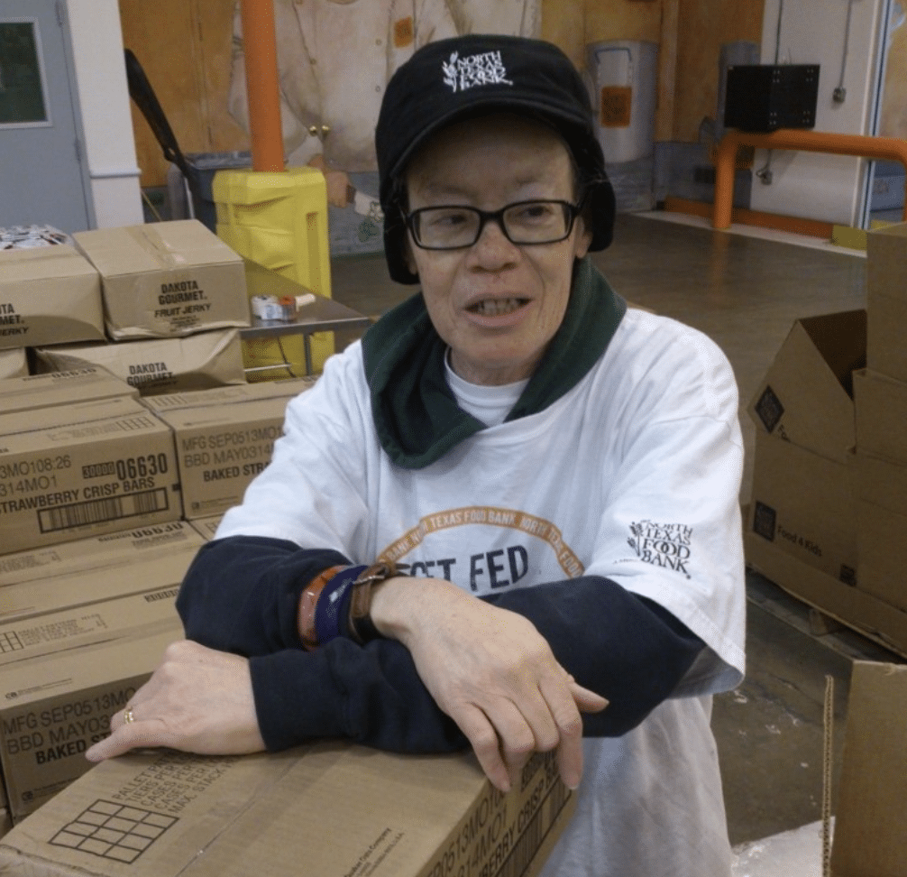 Older woman standing amongst boxes with an North Texas Food Bank hat