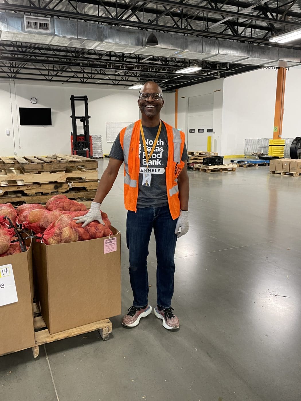 Many with orange safety vest smiling in warehouse next to box of potatoes