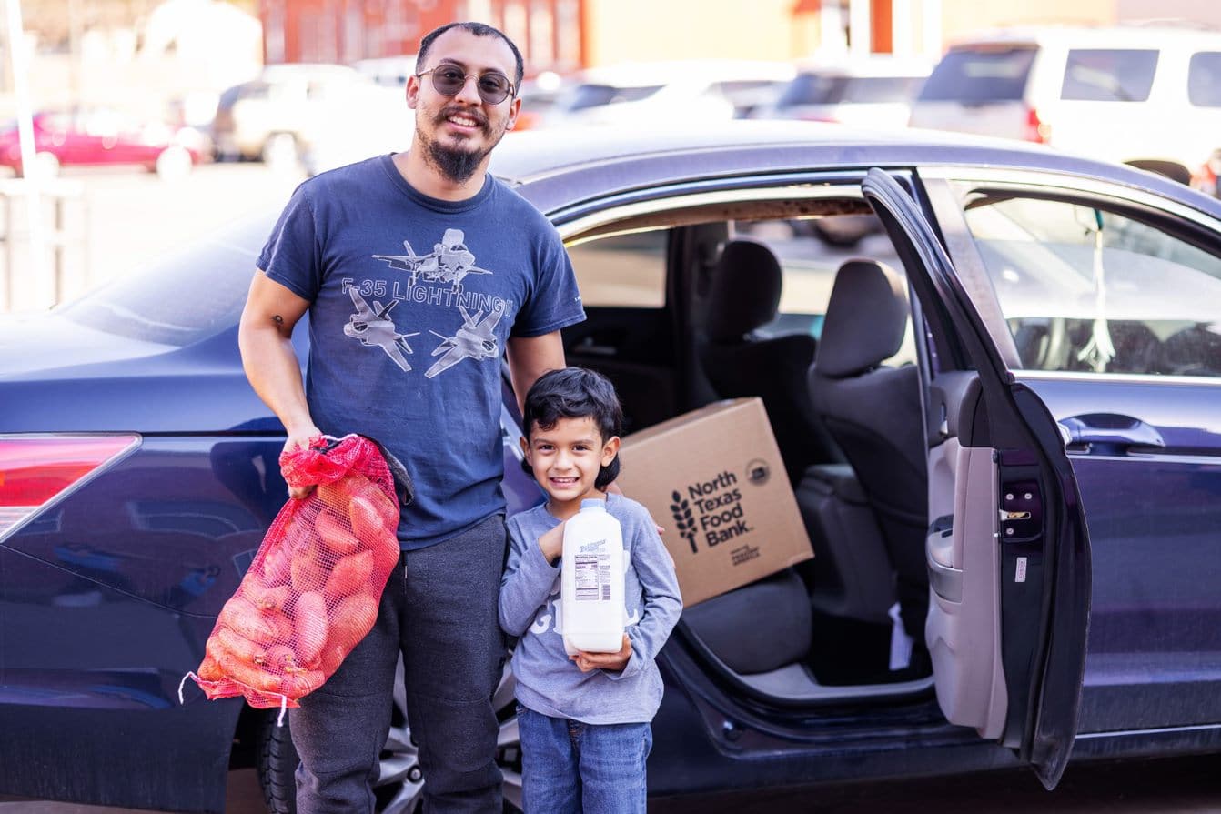 Father and son in front of car holding bag of potatoes and milk carton.