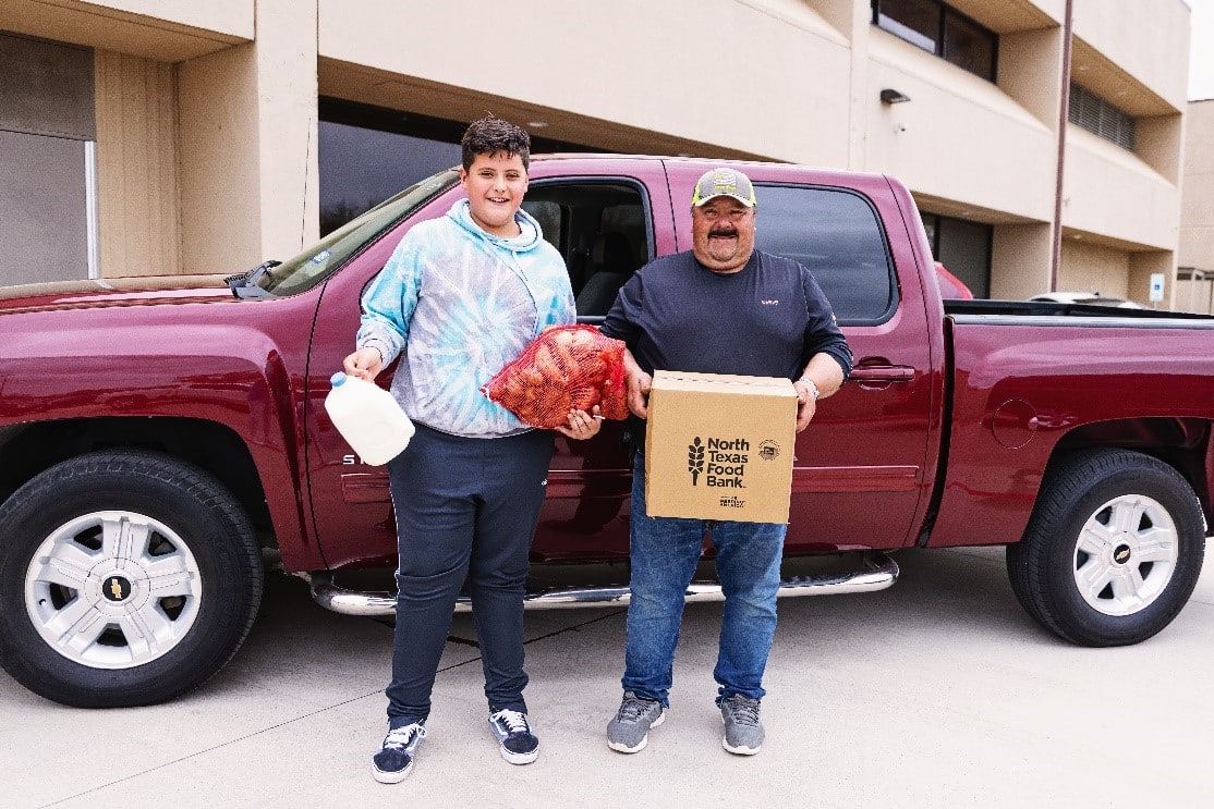 A father and son holding milk, potatoes and a box standing in front of a red pick-up truck.