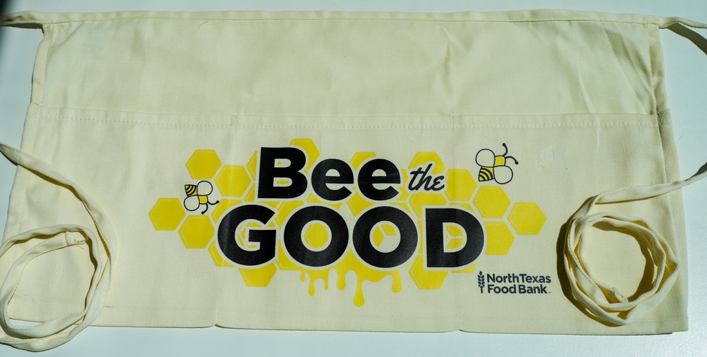Beige apron with Bee the Good in black text at the North Texas Food Bank store.
