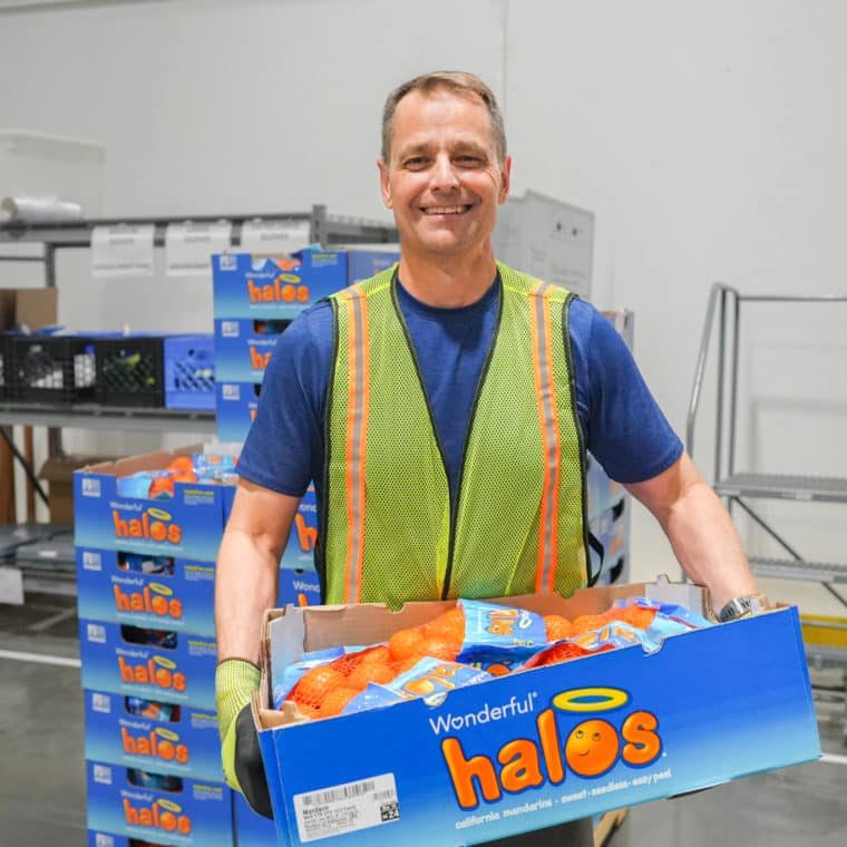 Smiling man in a hi-visibility vest stacks boxes of Halo mandarins in a warehouse margin area.