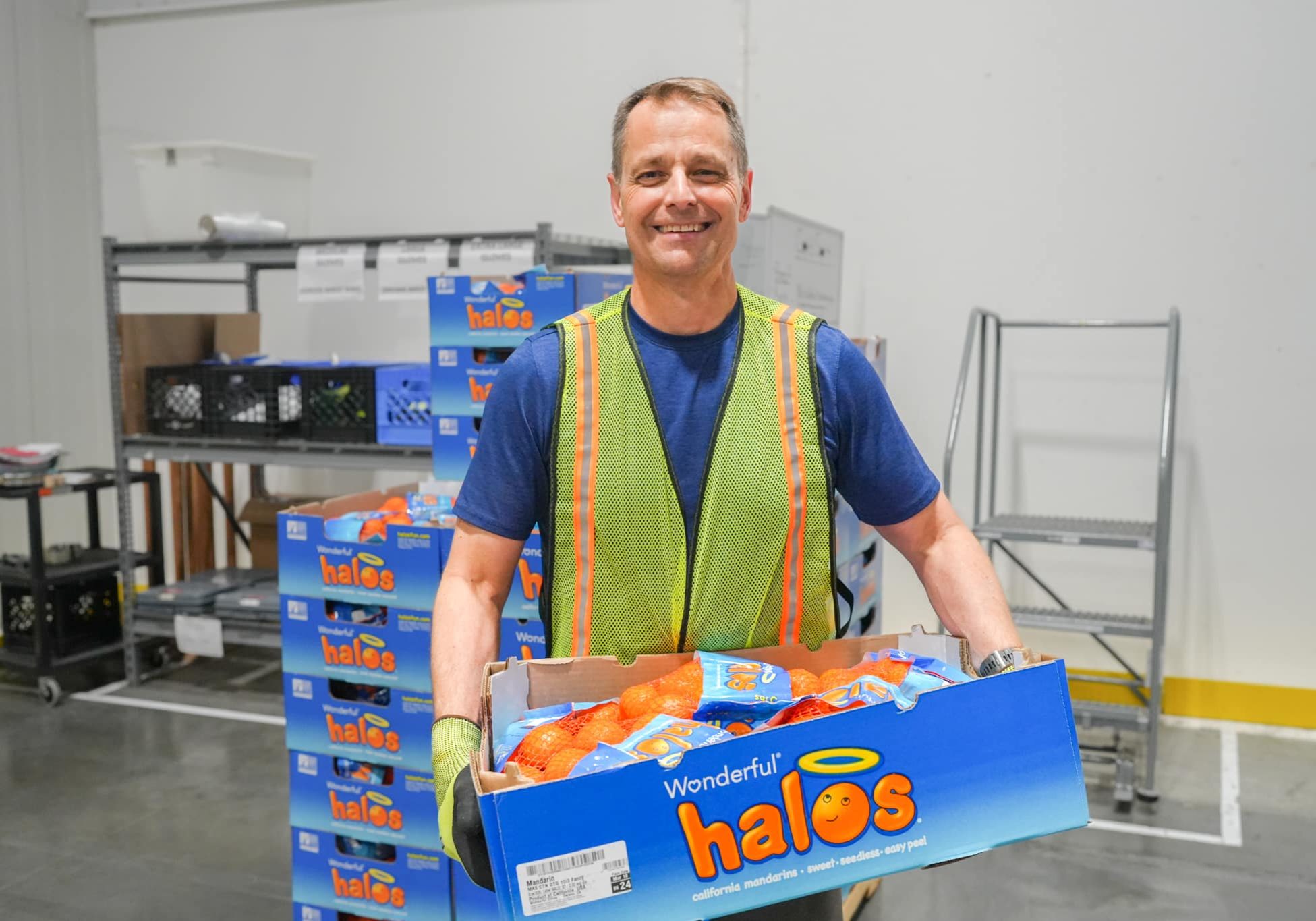 Smiling man in a hi-visibility vest stacks boxes of Halo mandarins in a warehouse margin area.