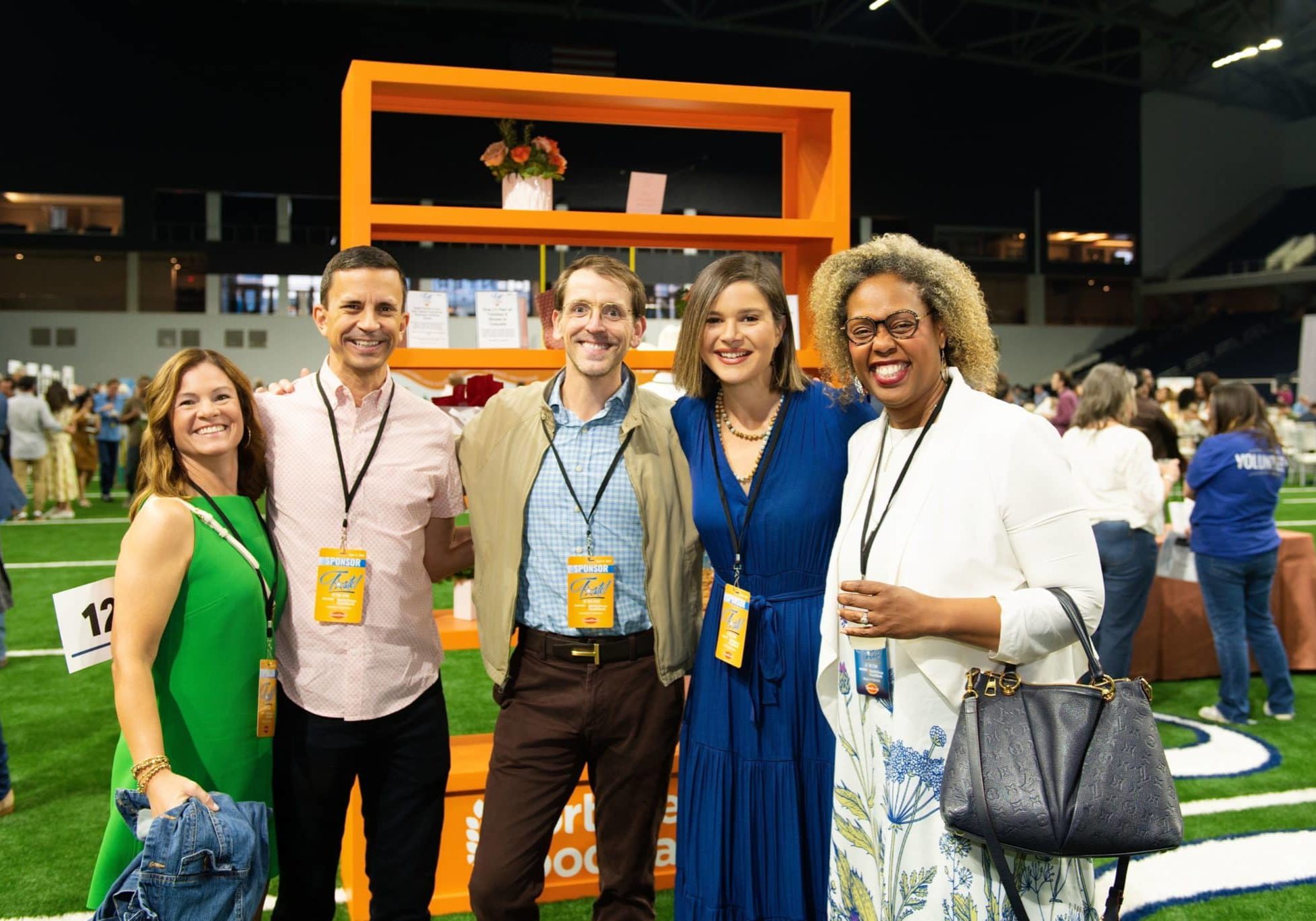 Five smiling adults posing for a photo at an indoor event, wearing sponsor badges with an orange display shelf behind them and a crowd in the background.