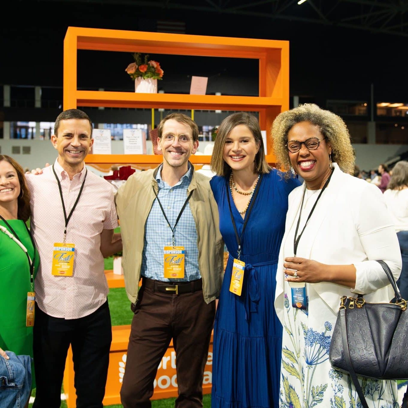 Five smiling adults posing for a photo at an indoor event, wearing sponsor badges with an orange display shelf behind them and a crowd in the background.