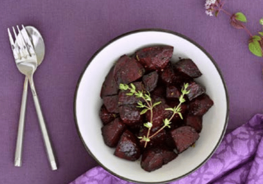 Roasted beets on a white plate with a sprg of rosemary on a purple tablecloth and a fork and knife
